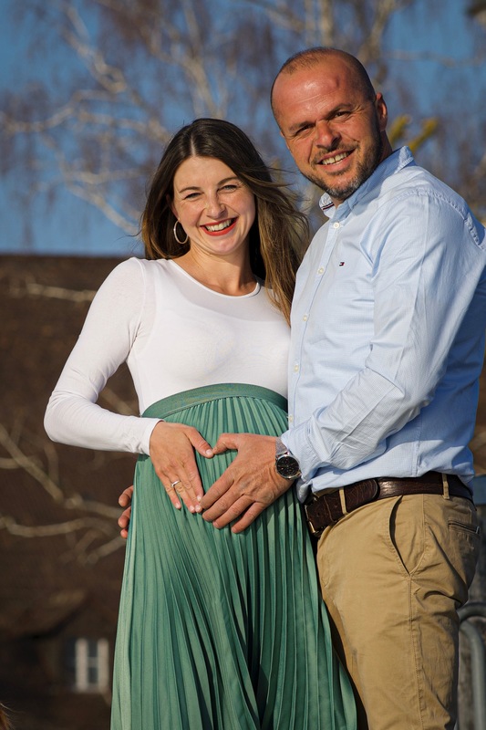 Babybauchshooting outdoor: glckliches Paar kurz vor der Geburt ihres zweiten Kindes, beide formen mit ihrer Hand ein gemeinsames Herz auf den Bauch